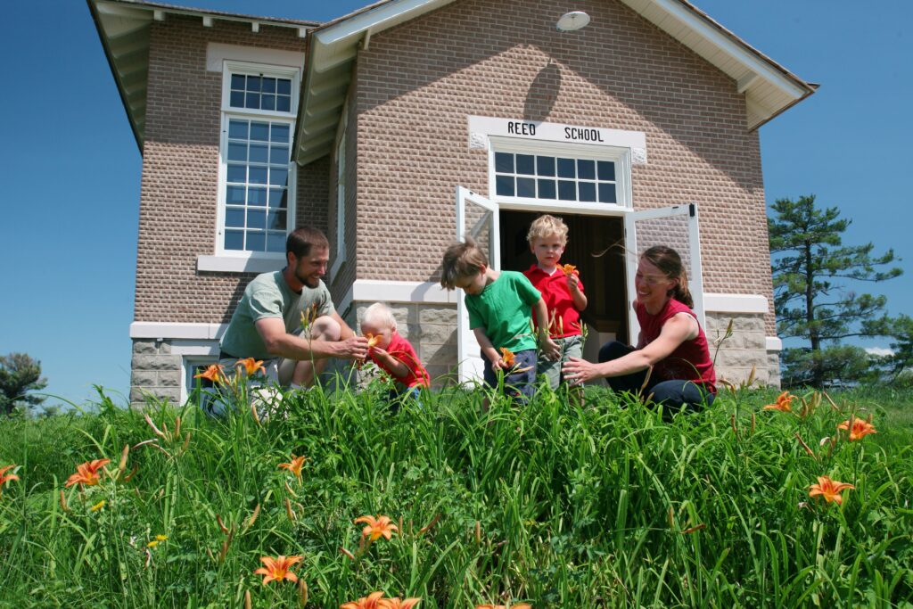 a family with three children play with the flowers in the grass in front of the Reed School building